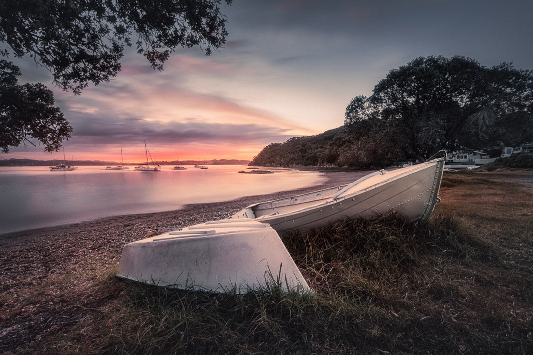 Photo of boats moored on a pebble beach framed by trees with the setting sun behind at Russell, Bay of Islands, New Zealand.