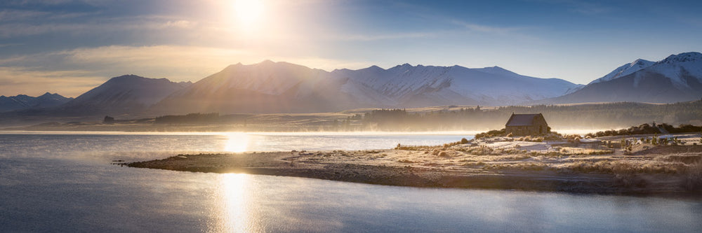 Panorama photo print of the sun rising up over Lake Tekapo with the Church of the Good Shepherd in the foreground. Snow is on the ground and surrounding mountains.