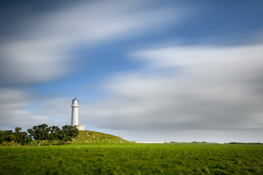 Photograph of Cape Egmont lighthouse with green grass and fields in the foreground and blue sky and white clouds in the sky