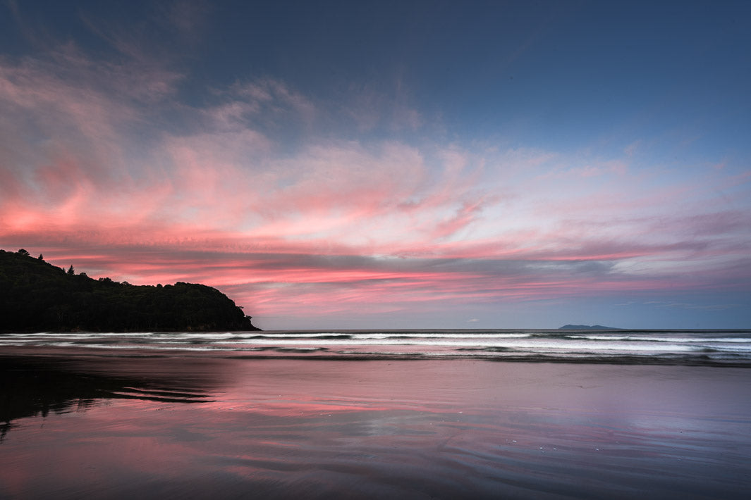 Sunrise pink clouds and blue skies reflected in the sea ripples over Waihi Beach