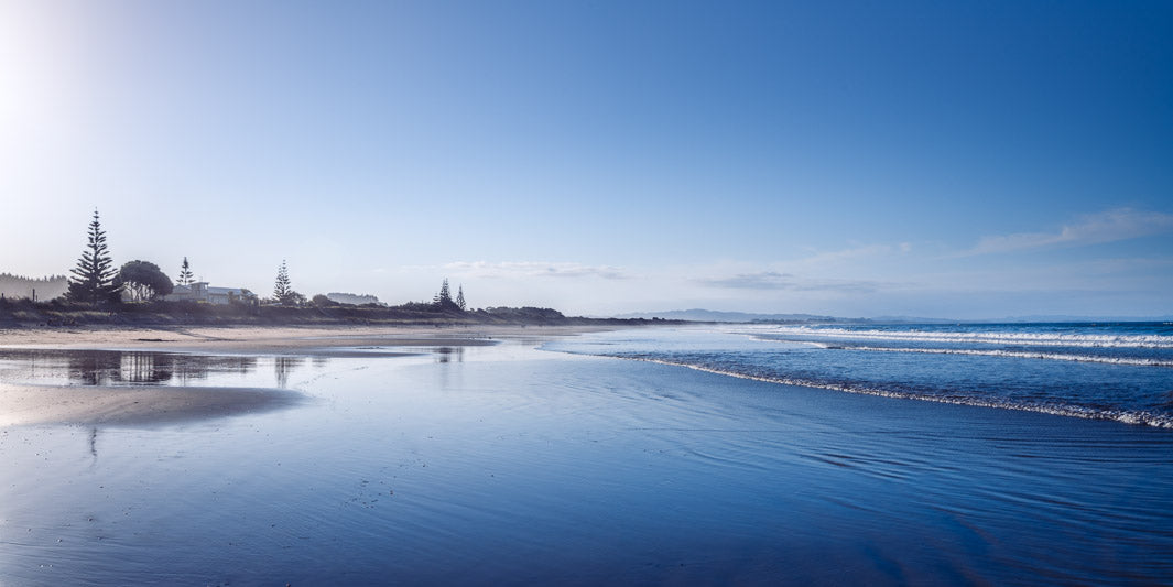 Landscape panorama photo of blue sky, blue sea, sand and trees in the distance at Waipu Cove in New Zealand.